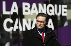 A man in glasses in an overcoat and red tie frowns in front of a La Banque du Canada sign.