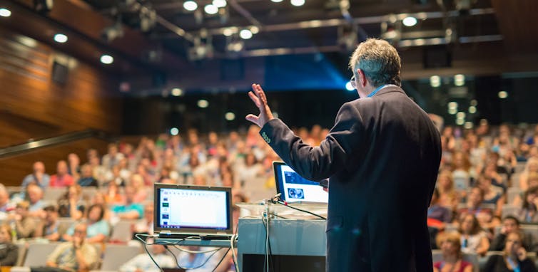 Man stands in front of large hall where he is lecturing hundreds of students