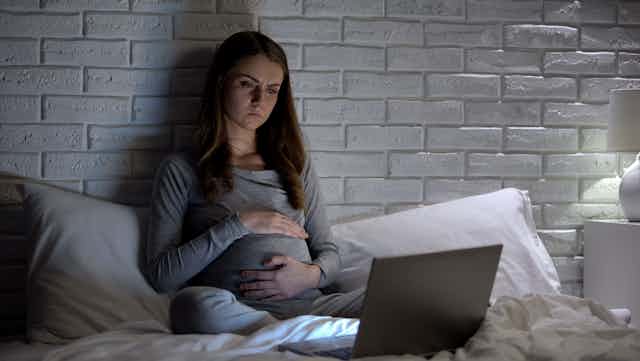 A young pregnant woman sits in bed looking at her laptop screen.