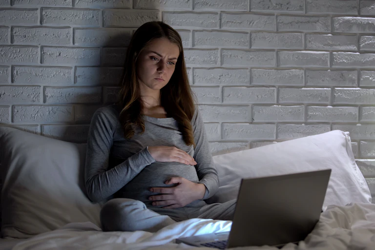 A young pregnant woman sits in bed looking at her laptop screen.