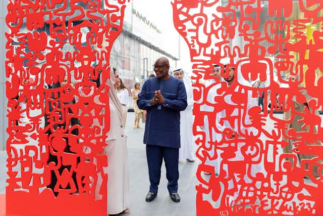 A man in Nigerian attire holds his hands in front of him and smiles at someone next to him. He is seen through a gap in a large red sculptural screen comprised of symbols.
