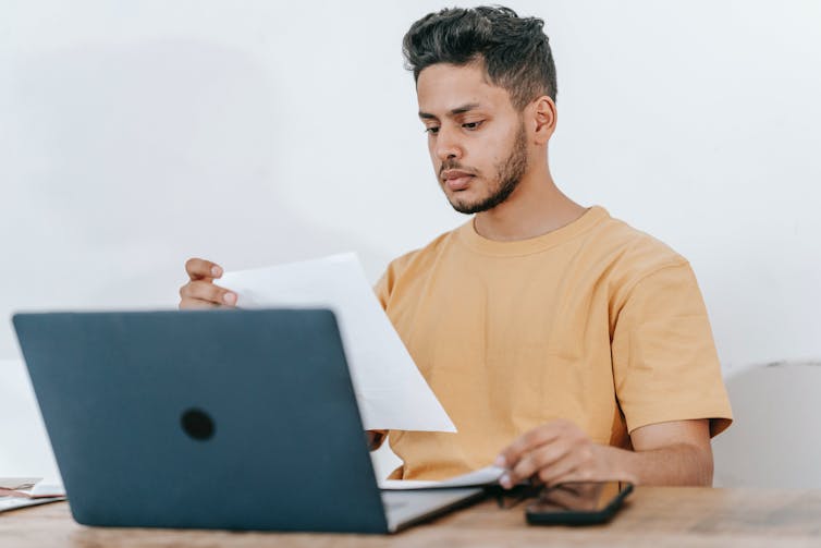 A young man reads a sheet of paper while sitting at a desk, with a laptop.