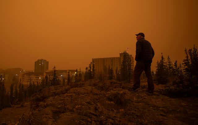 A man stands on a rocky hill overlooking buildings against an orange smoky sky.