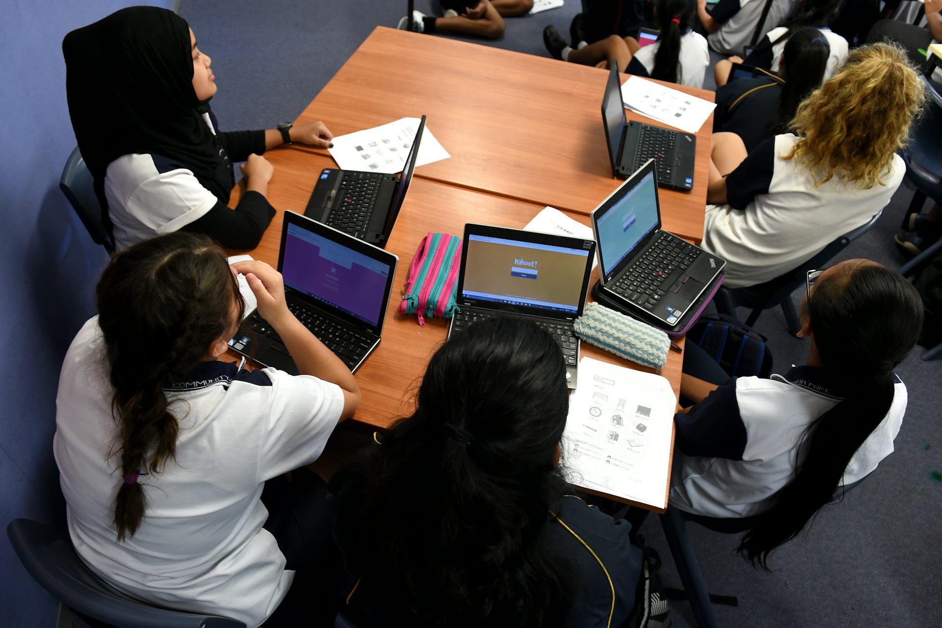 A classroom table with four students seated with laptops, listening to their teacher.
