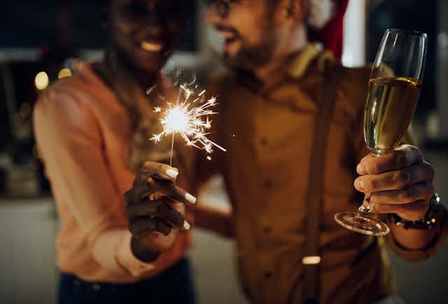 Close up photo of a smiling couple at a new year's eve party, the woman is holding a sparkler and the man is holding a glass of champagne