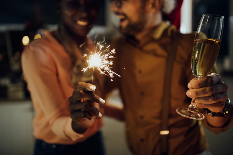 Close up photo of a smiling couple at a new year's eve party, the woman is holding a sparkler and the man is holding a glass of champagne