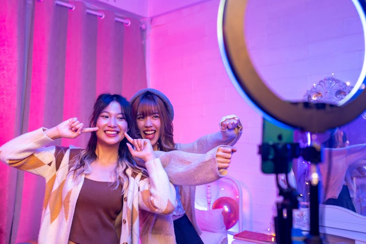 Two young women recording a video for social media. They are smiling and posing in a pink-lit bedroom, in front of a ring light and smartphone on a tripod.