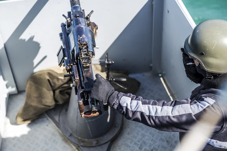 A gloved hand resting on the muzzle of a machine gun facing out into a body of water