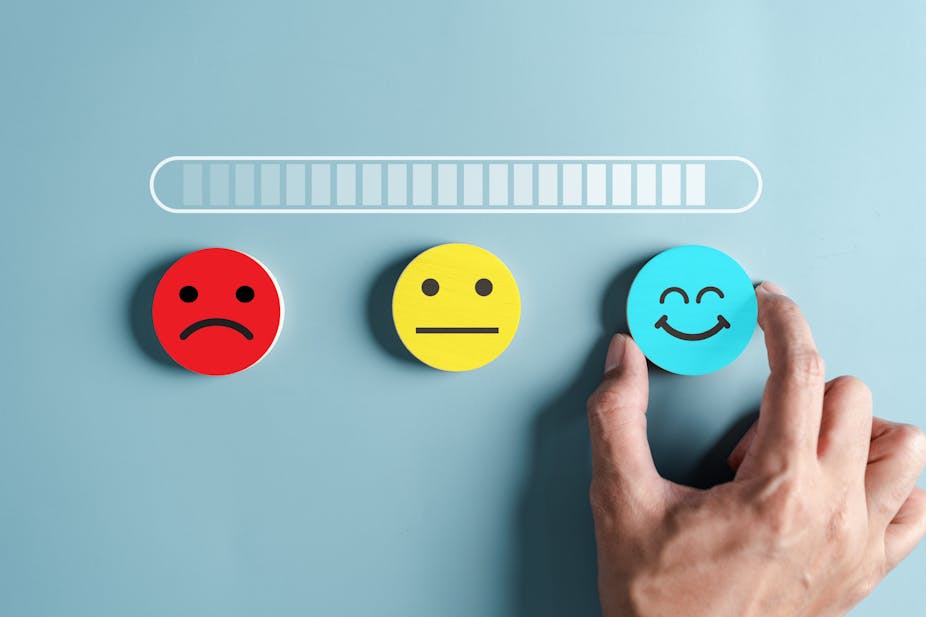 Three emoji faces – angry, no expression, and happy. A person chooses the happy emoji with their hand.