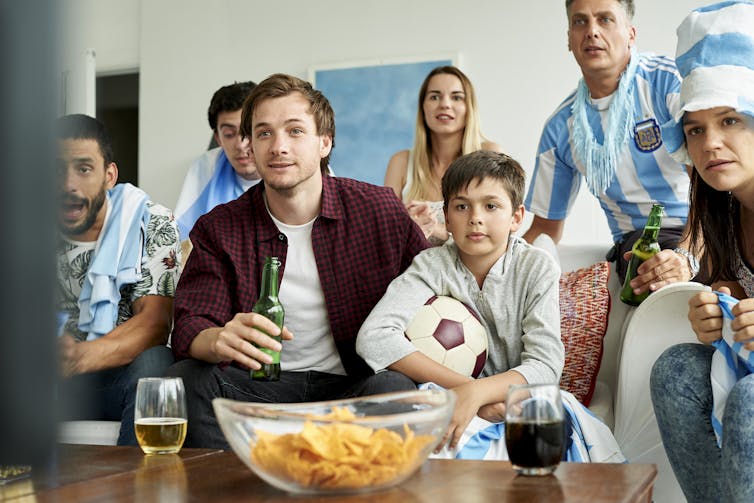 family, some with beers, watching a soccer match on TV, boy holds ball