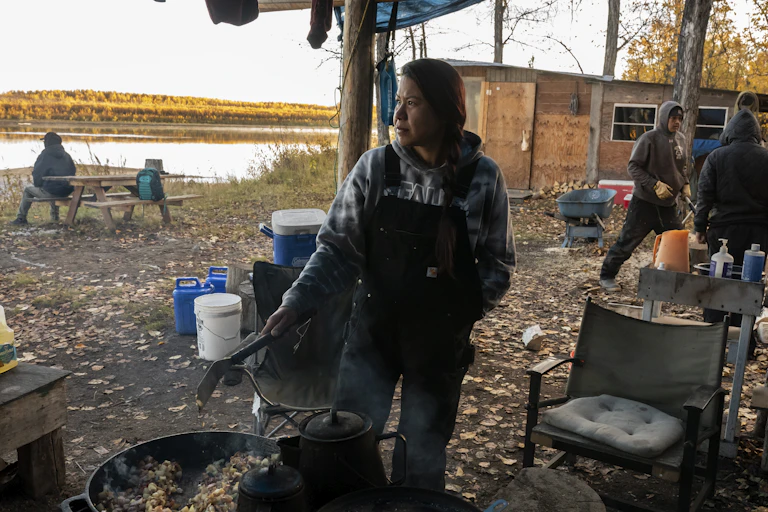 Hunters at camp along a river. One person is making food, others are talking or sitting at a picnic table.