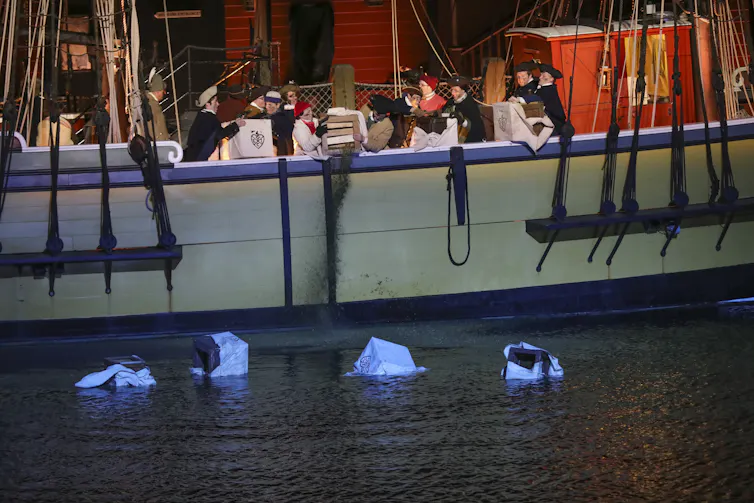 People dressed as colonists stand at a ship's rail and throw boxes overboard and empty tea into the water.