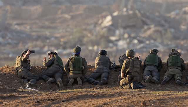 A line of soldiers in combat fatigues lie along a ridge looking over a community. 
