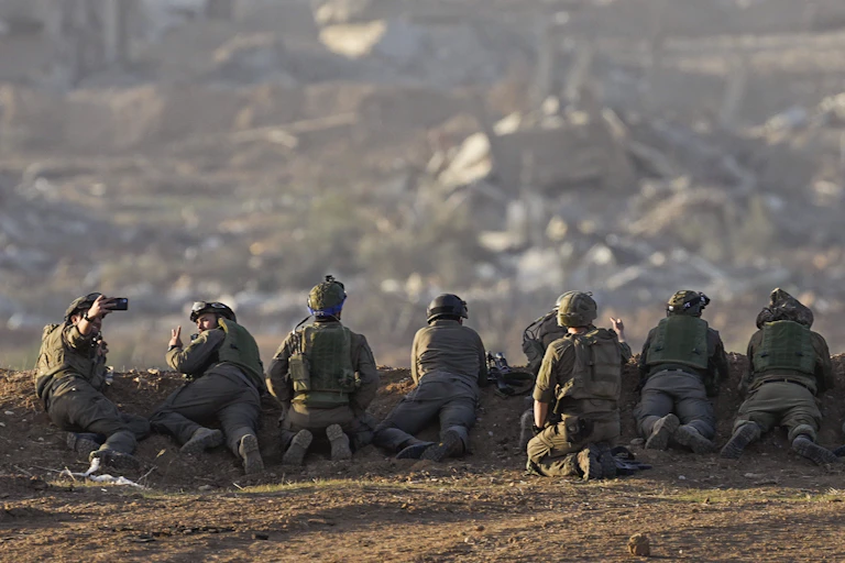 A line of soldiers in combat fatigues lie along a ridge looking over a community.