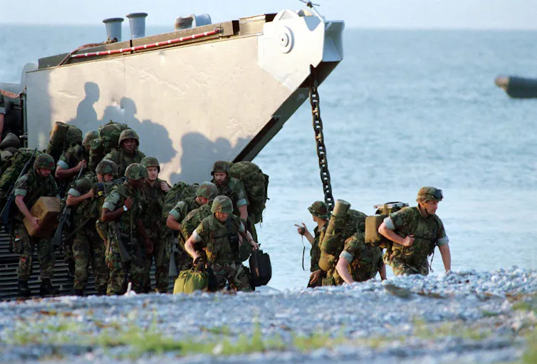 Soldiers in battle fatigues in a trench get off a boat on a beach.