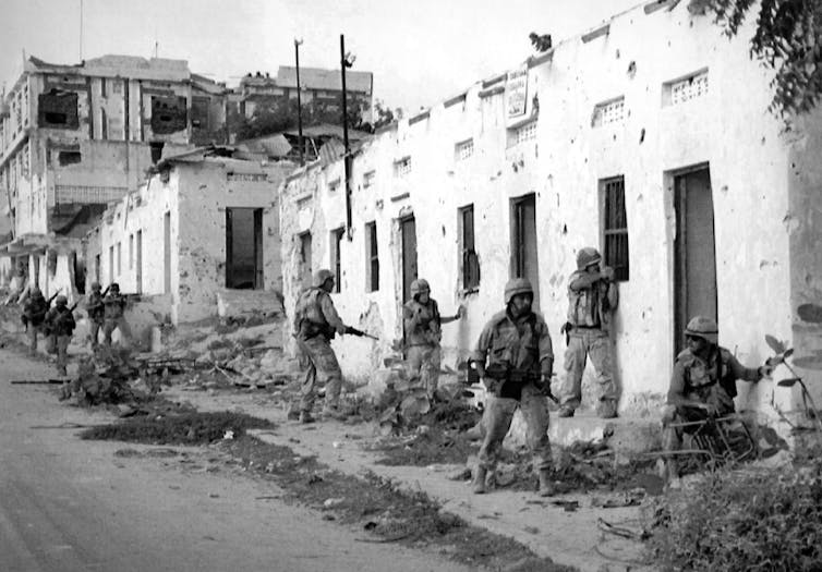 A black-and-white photo shows heavily armed soldiers looking into a white building and watching a road.