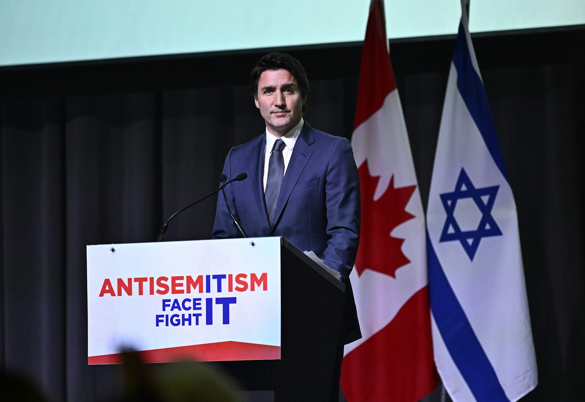 A dark-haired stands in front of a Canadian and Israeli flag and behind a podium with a banner that reads Antisemitism: Face it, fight it.