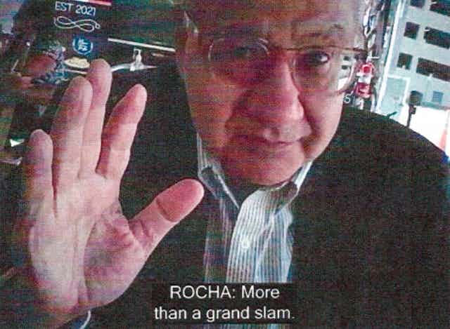 An elderly man facing a camera raises his hand.