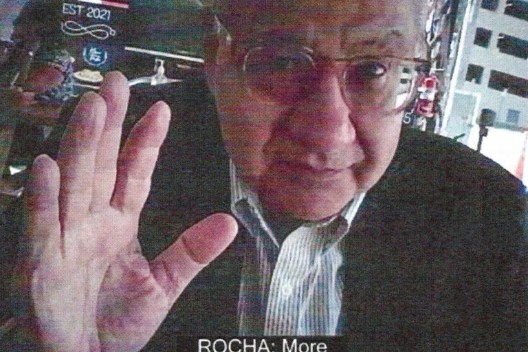 An elderly man facing a camera raises his hand.