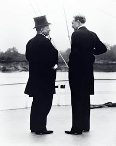 Two middleaged white men are talking with each other as they stand on boat that is traveling on a river.