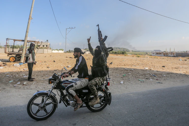 A person drives a motorcycle while a person holding a military-style rifle rides on the back.