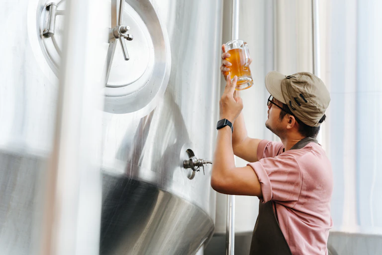 brewer in apron holds up beer stein next to tank