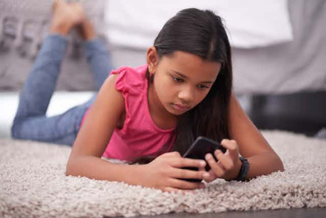 A young girl lies on a rug while looking at social media on her phone.