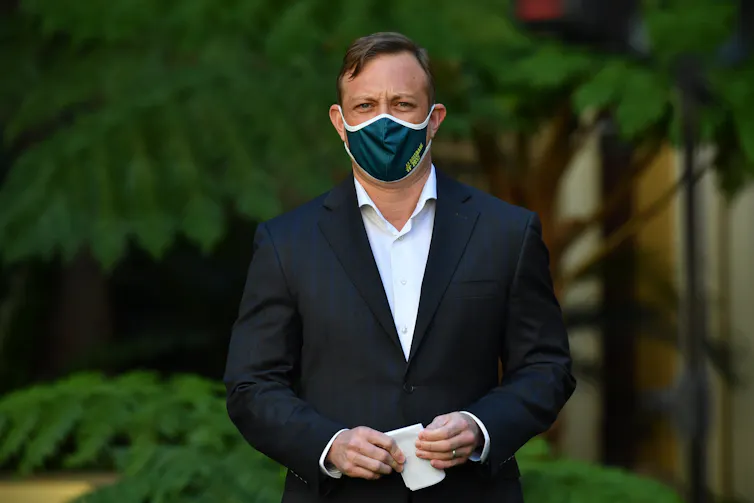 A man in a suit and a face mask stands in front of some plants
