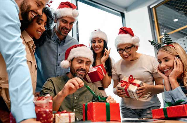 A man opening a gift, surrounded by colleagues in business attire and Santa hats.
