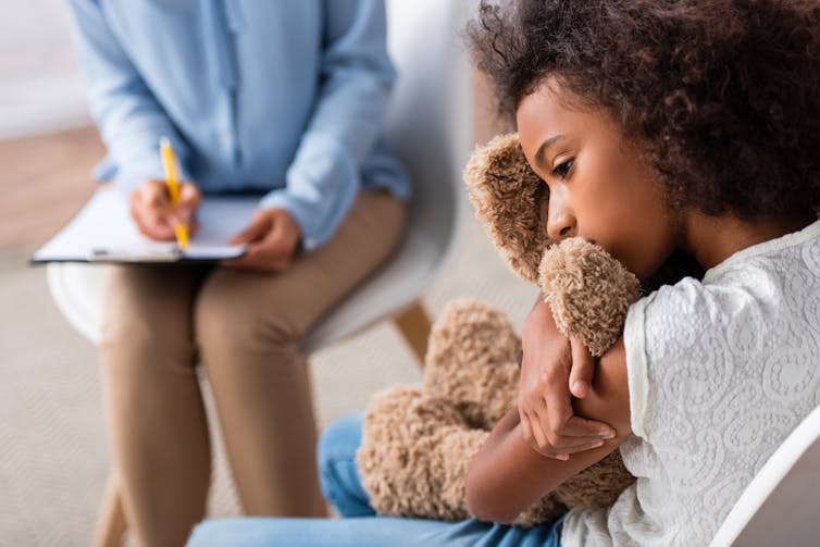 girl hugs teddy while adults sits nearby with clipboard