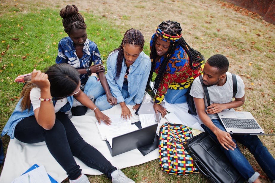 A group of five young people sitting on a blanket on some grass, crowded around a laptop and studying some papers