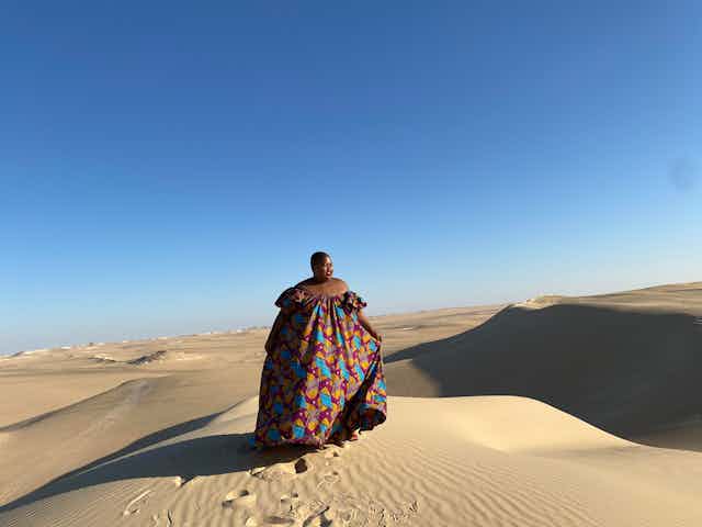 A blue sky and desert stands. An African woman with a shaved head poses for a photo on top of a sand dune. She wears a flowing African print dress.