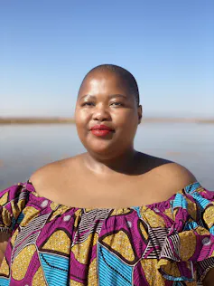 An African woman smiles slightly as she looks at the camera, her hair shaven and wearing African print.