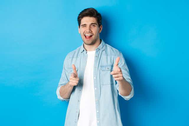 A man winking and pointing finger guns at the camera in a flirty way, standing against a blank blue wall