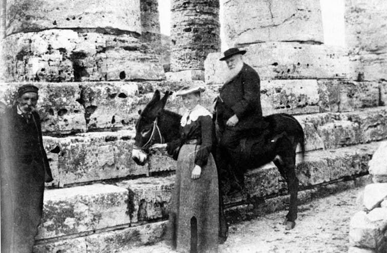 A Victorian black and white photo showing a man sitting on a donkey led by a woman with another man standing nearby.