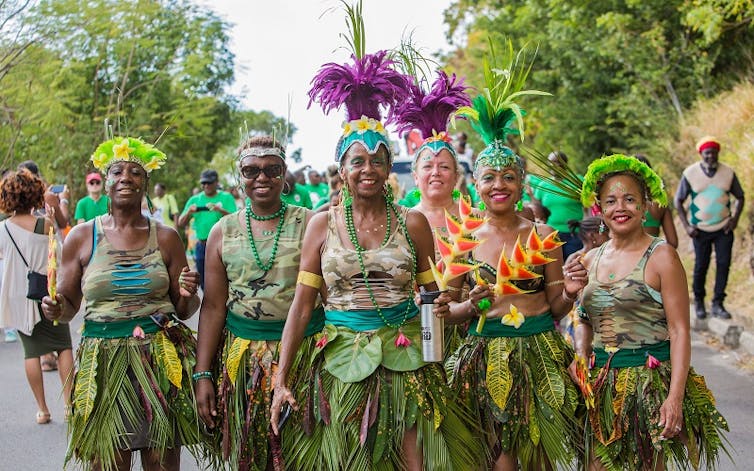 Group of mostly women of colour wearing colourful headdresses, green camouflage vests and green skirts made from palm leaves.