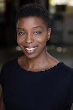 An elegant African woman with natural hair and a black blouse smiles at the camera.