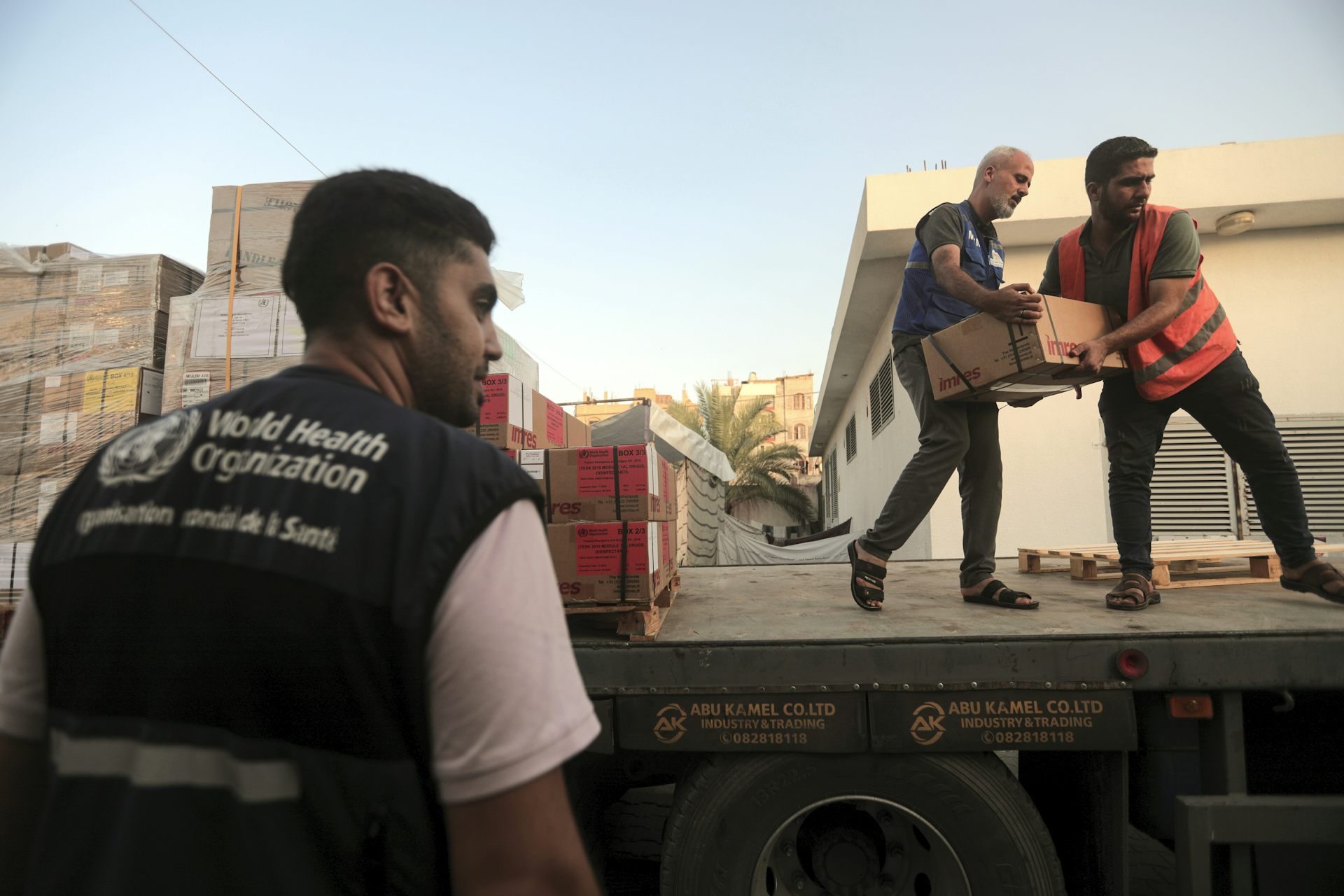 Deux hommes transportent des cartons depuis un camion, tandis qu'un troisième se tient sur le côté. Il porte un gilet de l'Organisation mondiale de la santé.