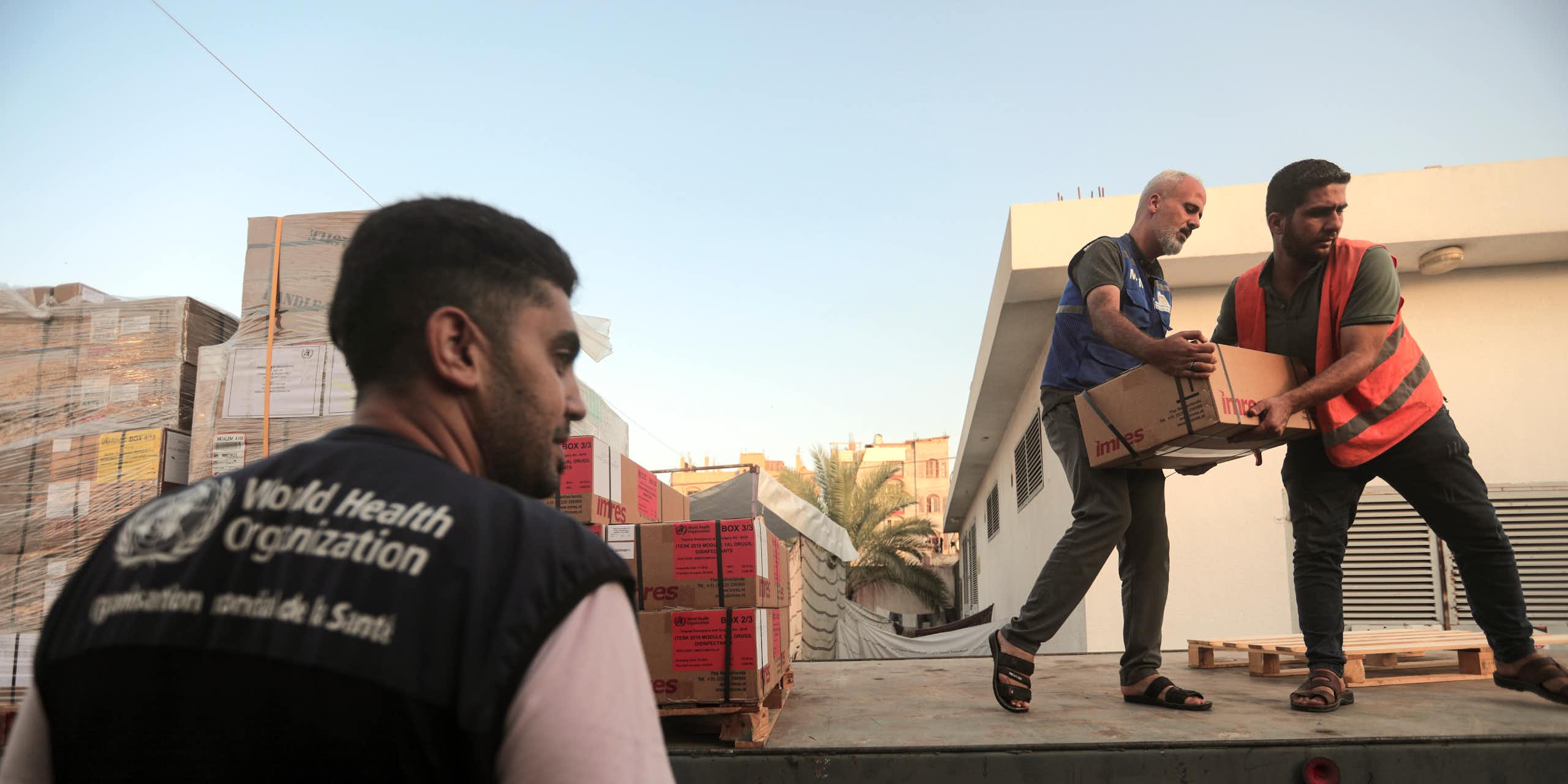 Deux hommes transportent des cartons depuis un camion, tandis qu'un troisième se tient sur le côté. Il porte un gilet de l'Organisation mondiale de la santé.