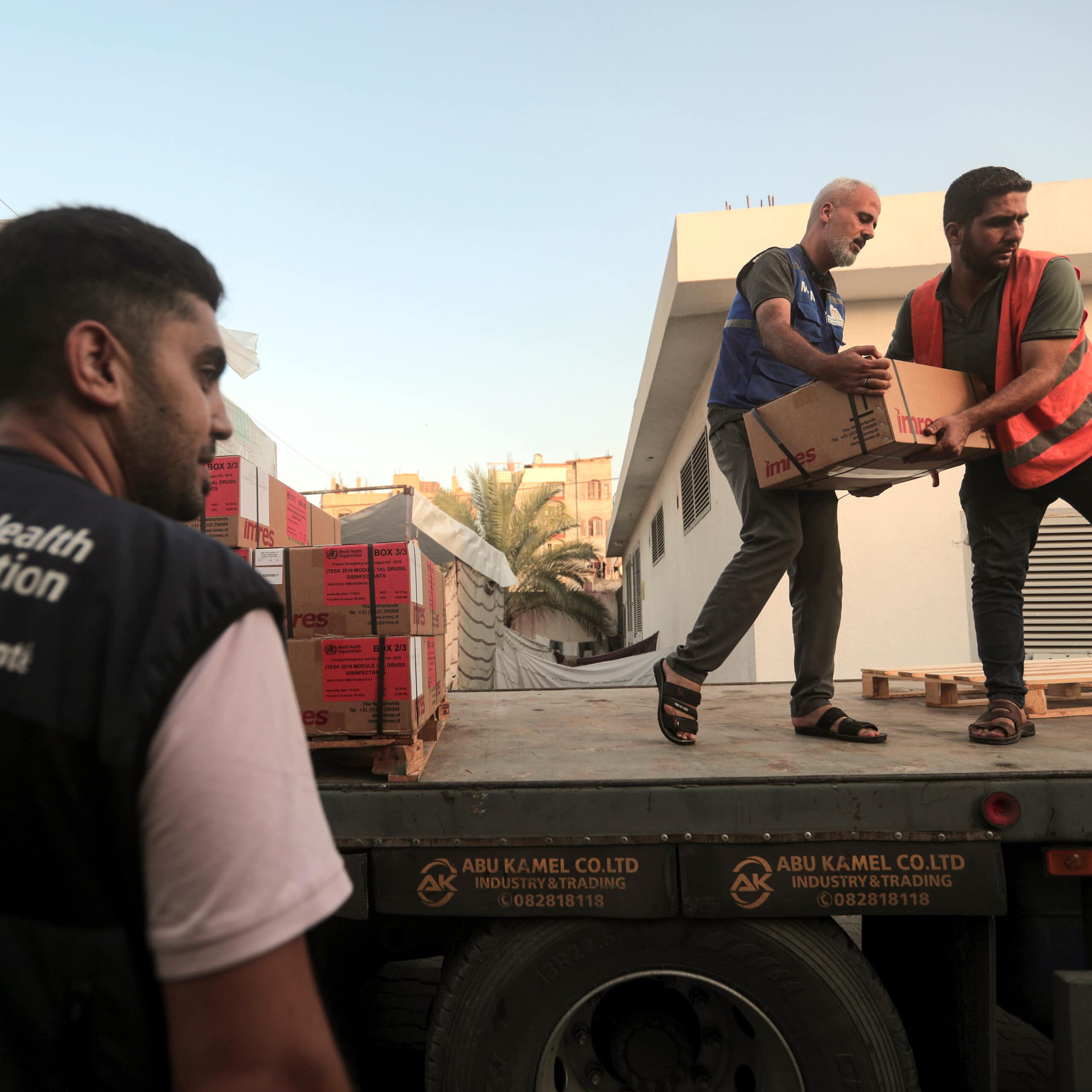 Deux hommes transportent des cartons depuis un camion, tandis qu'un troisième se tient sur le côté. Il porte un gilet de l'Organisation mondiale de la santé.