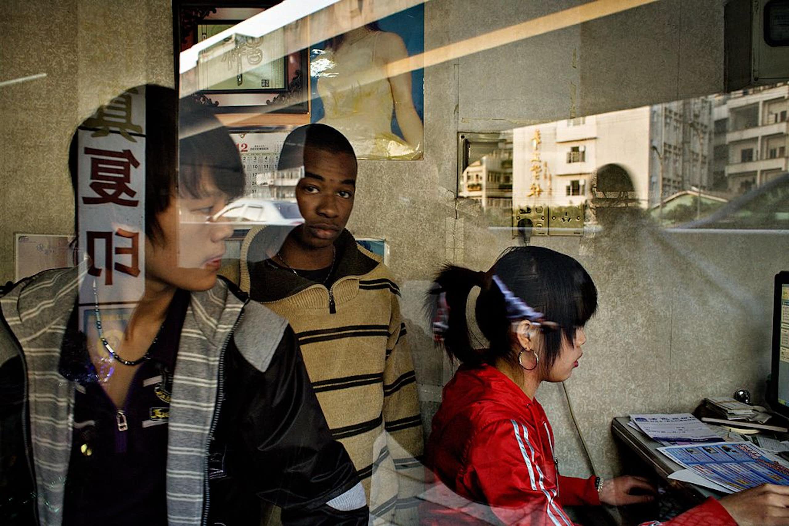 Through the glass of a shop front, three people are visible. A chinese woman and man flank an African man who looks into camera through the glass. A blurry reflection of a woman is seen passing on the street.