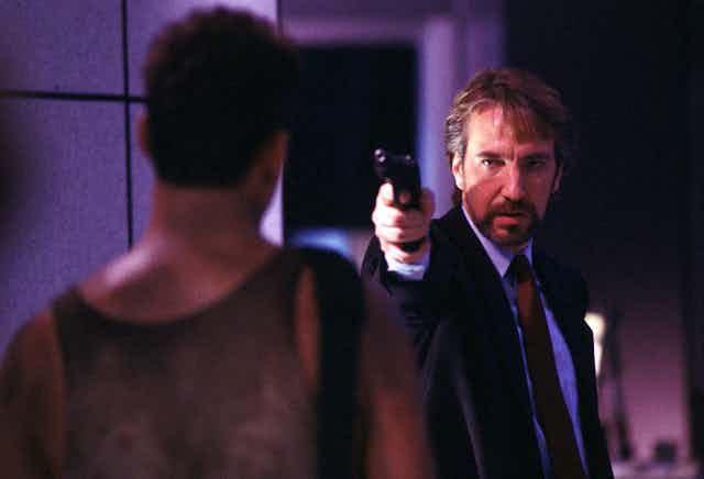 A still from the movie Die Hard, showing Alan Rickman as Hans Gruber holding a gun pointed at Bruce Willis