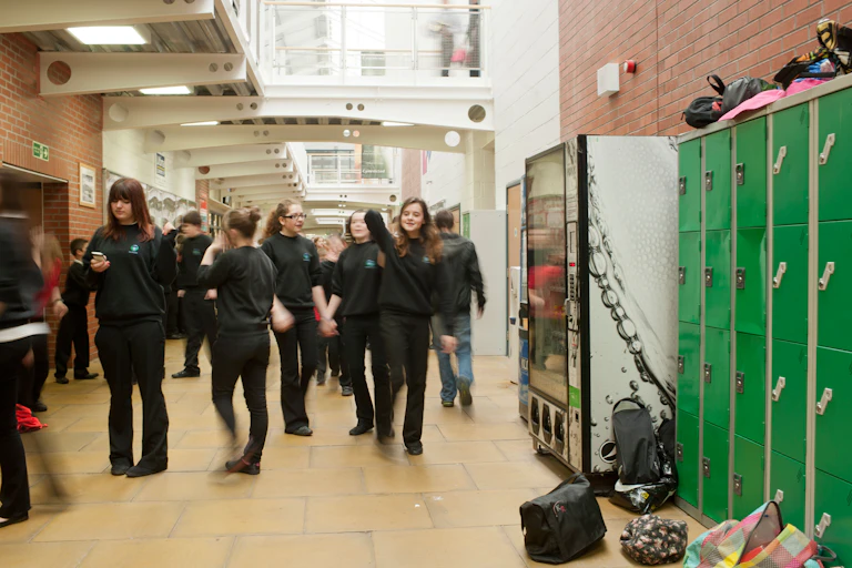 A group of teenagers in uniform stand in a school corridor next to green lockers.