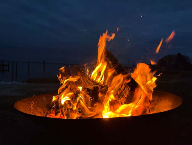 Two wooden logs burn up in a small campfire sitting in a metal firepit, against a navy blue sky.