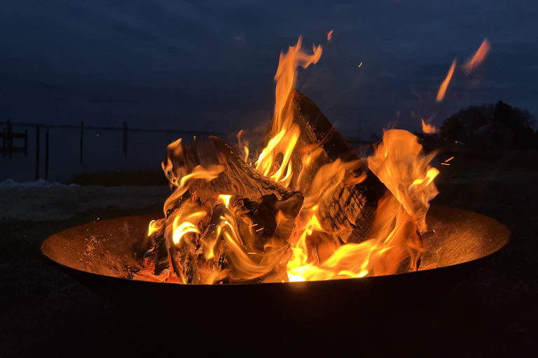 Two wooden logs burn up in a small campfire sitting in a metal firepit, against a navy blue sky.