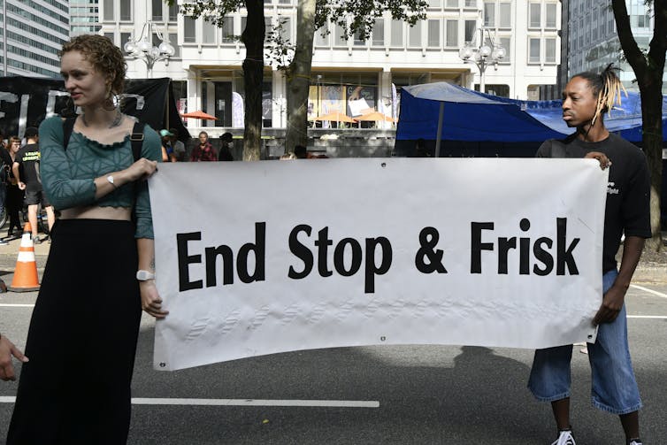 Dos jóvenes en una plaza urbana sostienen un cartel que dice 