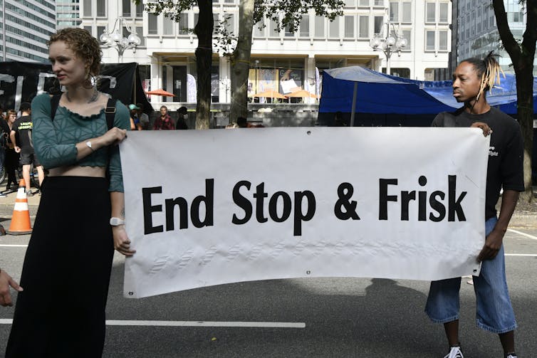 Two young people in an urban plaza hold up a sign that reads