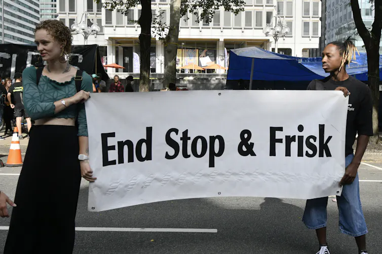 Two young people in an urban plaza hold up a sign that reads