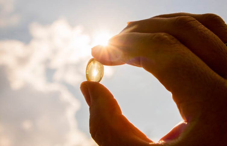 A person's hand holds a vitamin D supplement up towards a sunny sky.