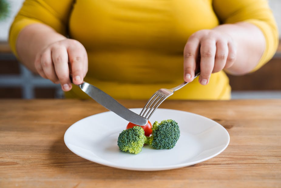 A woman uses a fork and knife to cut a piece of broccoli on her plate.